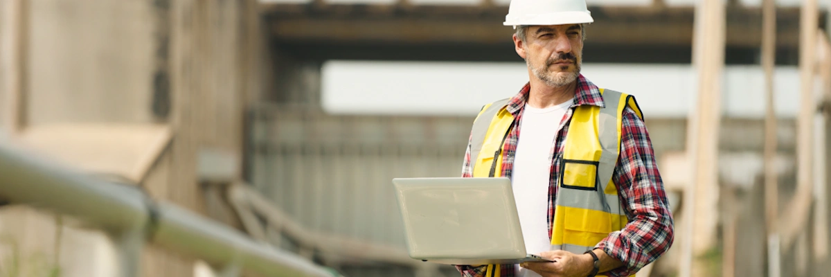 Construction manager in safety gear holding a laptop at an industrial site, monitoring project progress.