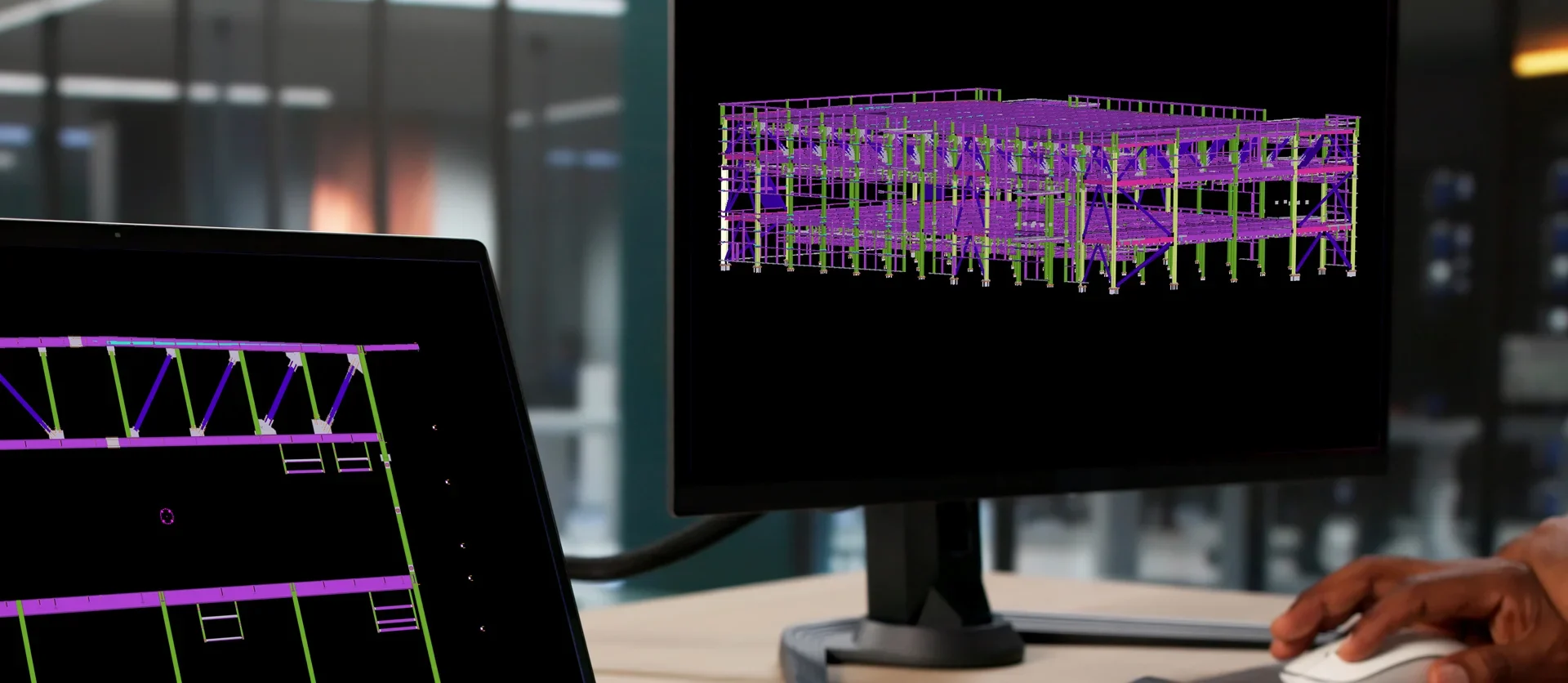A close-up of a user's hand using a computer mouse at a desk, with the main desktop monitor showcasing a complex, fully completed purple 3D structural matrix model of a multi-story building against a black background.