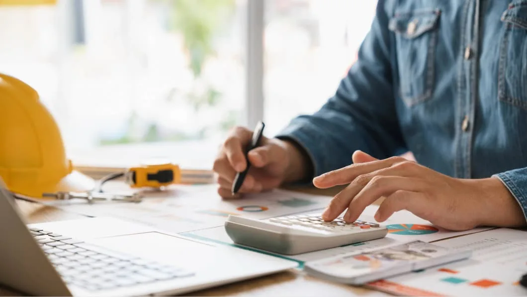 Close-up of a person using a calculator over blueprints and financial charts with a yellow hard hat nearby.