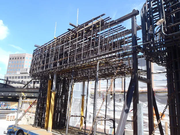 A large, complex cage of rebar, ready for concrete, is shown on a construction site with a modern building in the background.