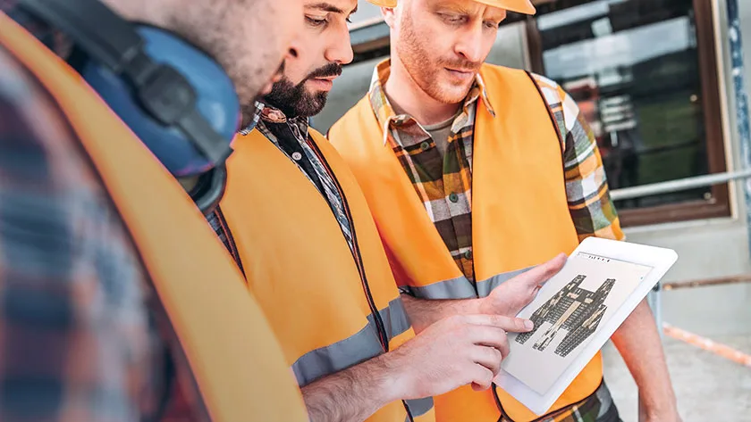 Three construction workers in hard hats and safety vests review a blueprint on a tablet.