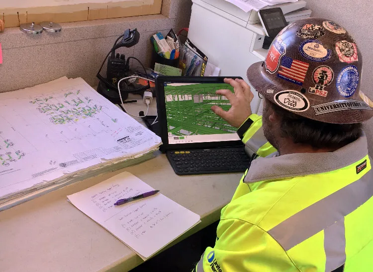 A construction worker in a high-visibility vest and a hard hat covered in stickers looks at a tablet displaying a 3D model of a building.