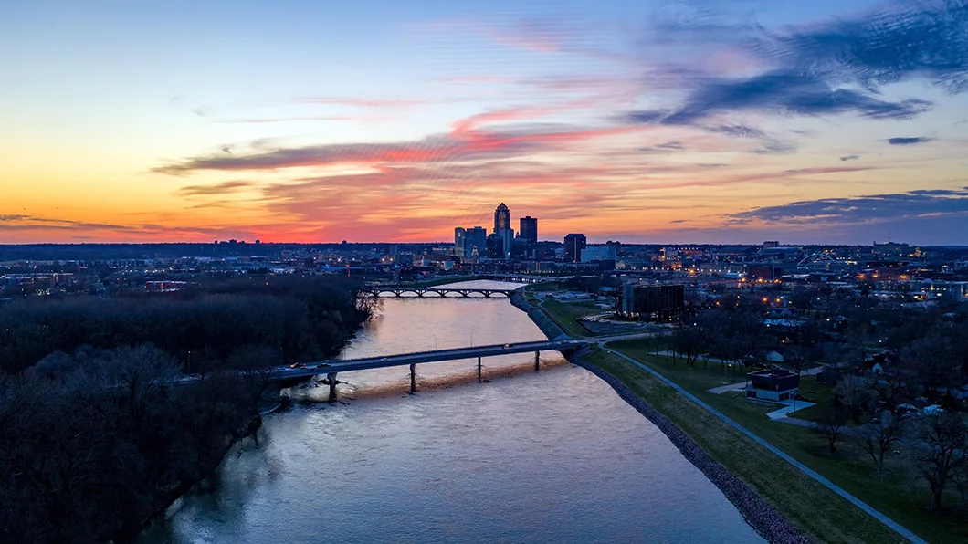 Aerial sunset over Des Moines, IA: cityscape, Des Moines River, and modern concrete-beam bridges, like the Grand Avenue Bridge.