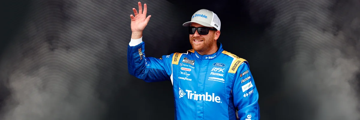 Chris Buescher, RFK Racing driver in a blue Trimble-branded suit and hat waves in front of a dark, smoky background with white geometric lines.