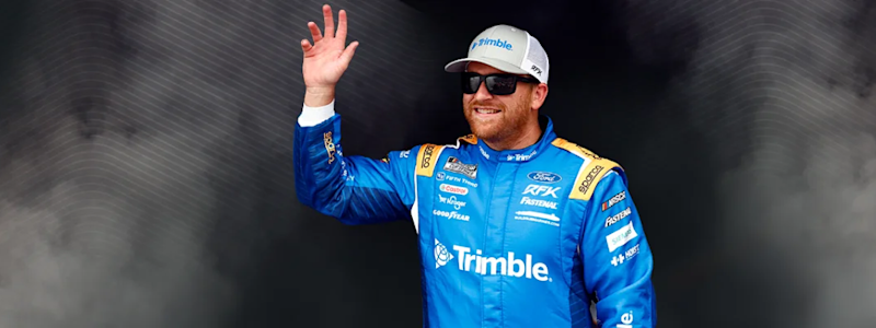 Chris Buescher, RFK Racing driver in a blue Trimble-branded suit and hat waves in front of a dark, smoky background with white geometric lines.
