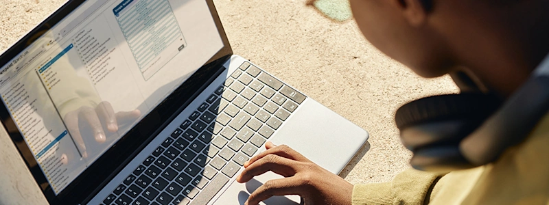 Student with headphones using a laptop outdoors in sunlight.