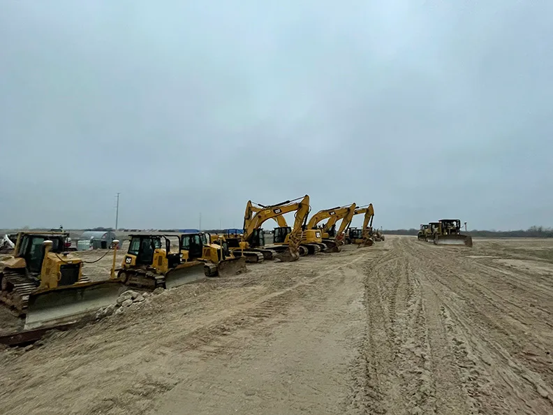 Yellow excavators and bulldozers are parked in a line on a dirt construction site under a cloudy sky.