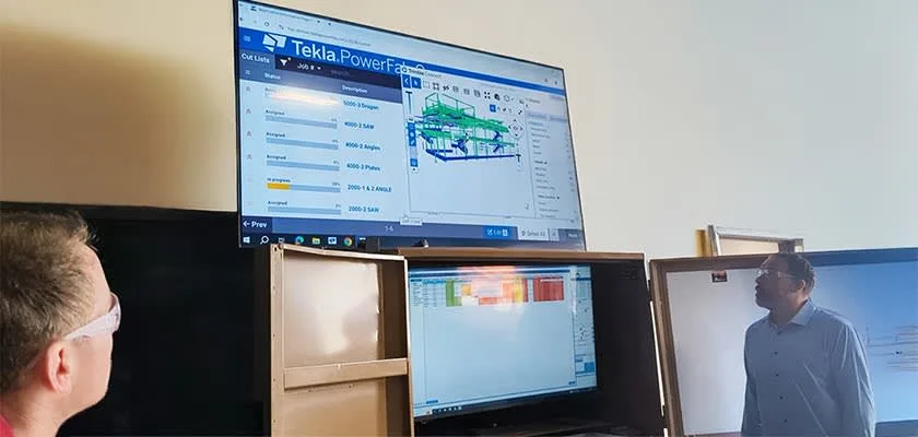 Two men look at a computer screen displaying Tekla PowerFab software with 3D structural modeling.