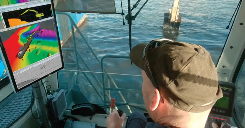 Operator controls a dredge from a cabin, viewing a 3D map of the seabed and the dredge's model, with the machine in water.