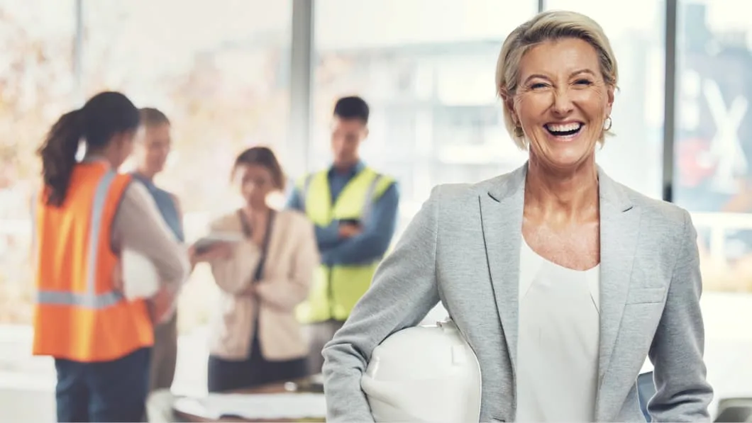 A smiling woman in a grey blazer holds a white hard hat in an office with blurred colleagues.