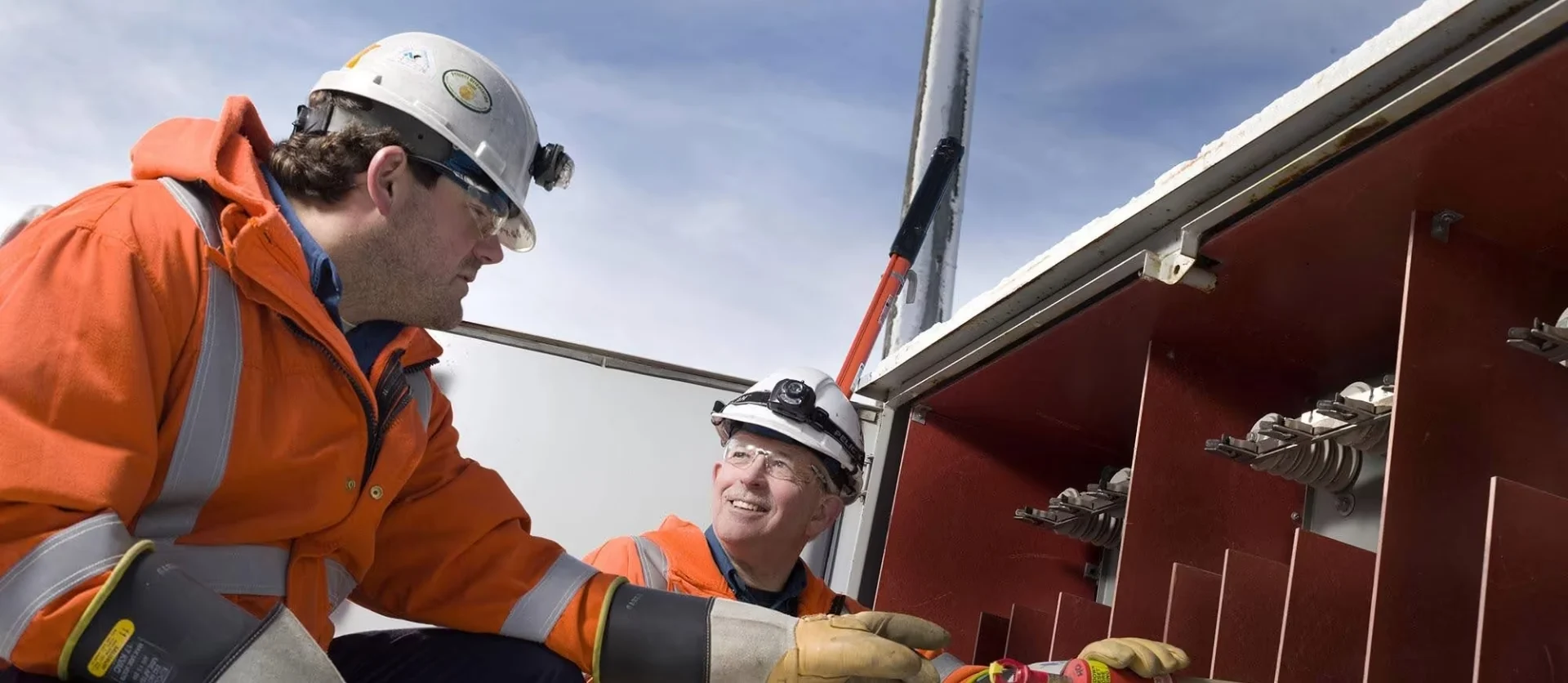 Two EPCOR workers in orange high-visibility gear and hard hats inspecting an open electrical cabinet.