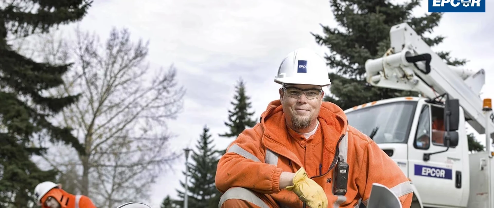 EPCOR utility worker in orange safety gear and hard hat kneeling by equipment with a bucket truck behind him.
