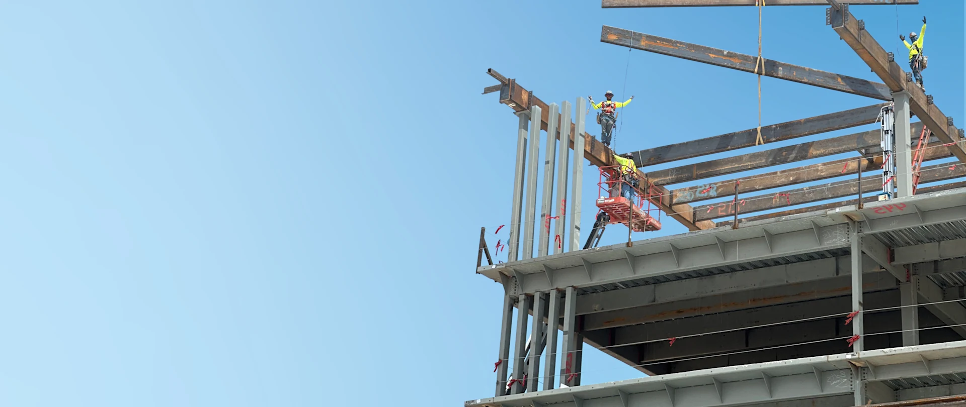 Construction workers in high-visibility gear and safety harnesses navigate a high-altitude steel framework under a clear blue sky.