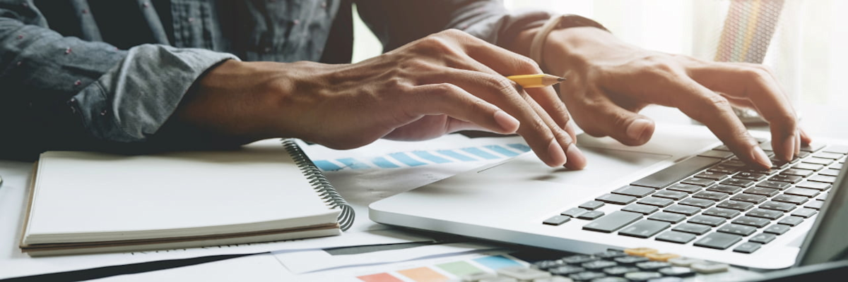 A person types on a laptop at a desk with a notebook, calculator and charts.