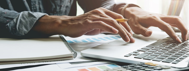 A person types on a laptop at a desk with a notebook, calculator and charts.