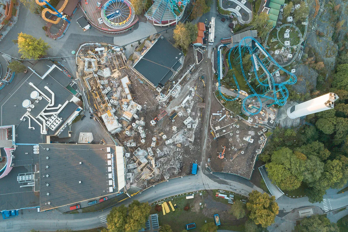 Aerial view of an amusement park under construction, showing a blue roller coaster, construction equipment, and building foundations.