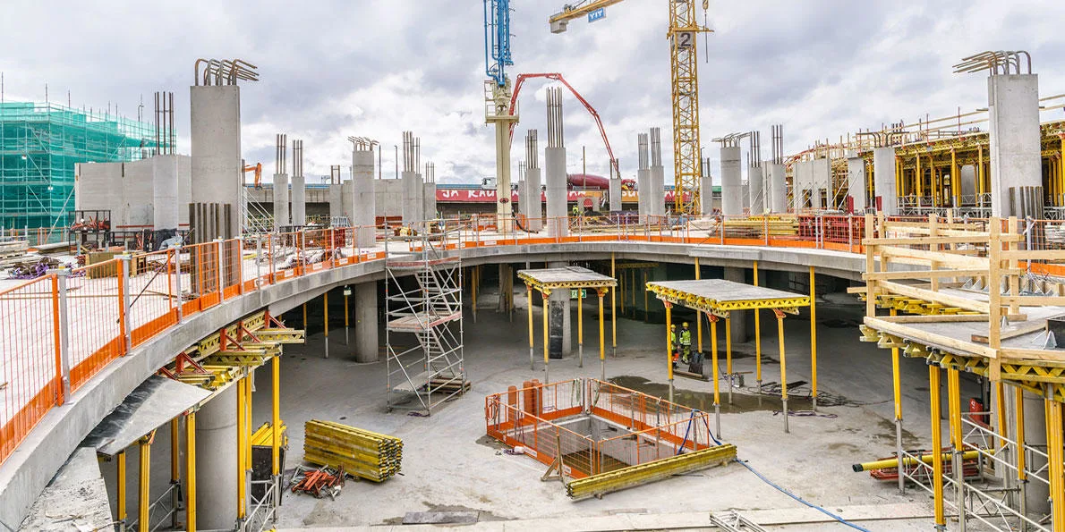 Construction site with concrete pillars, scaffolding, and cranes under a cloudy sky.