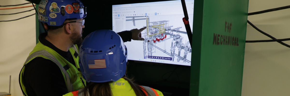 F+F Mechanical workers in blue hard hats and yellow vests review a 3D mechanical piping model on a large screen.