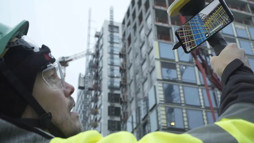 A construction worker in a hard hat and safety glasses holds a device with a smartphone displaying a building's blueprint.
