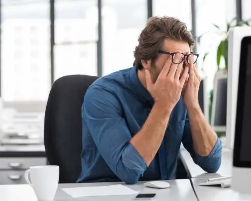 Main in blue button down shirt rubbing eyes under glasses in office setting.