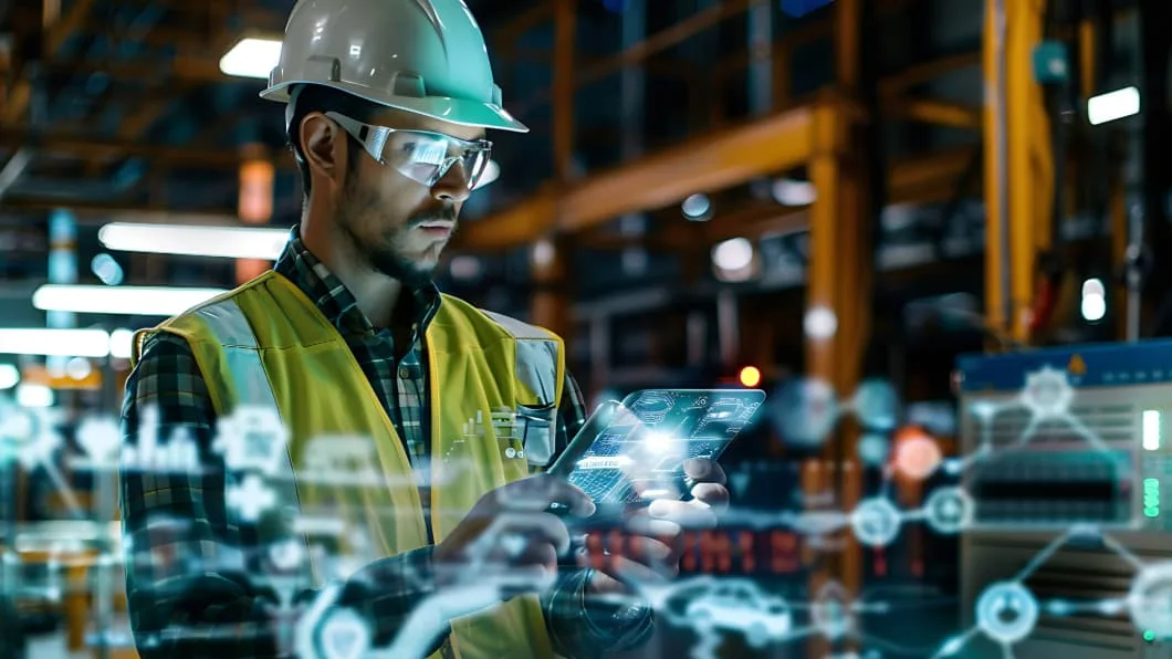 Worker in safety gear using a digital tablet with augmented reality data overlays in a factory.