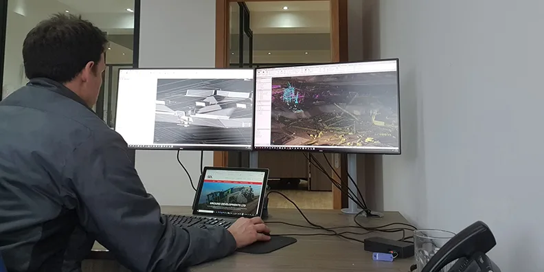 Architect works on dual monitors displaying 3D models and site scan data.