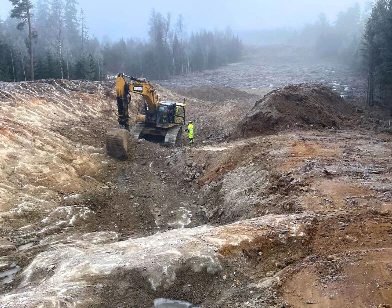 Excavator and worker in a muddy, wooded construction site on a foggy day.