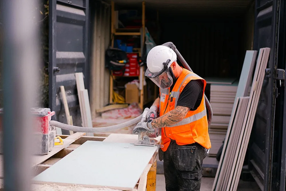 Construction worker in orange safety vest cutting material with power tool at worksite, wearing protective helmet and mask.