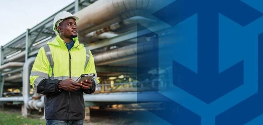 Engineer in hard hat and hi-vis jacket holding tablet in front of industrial pipes.