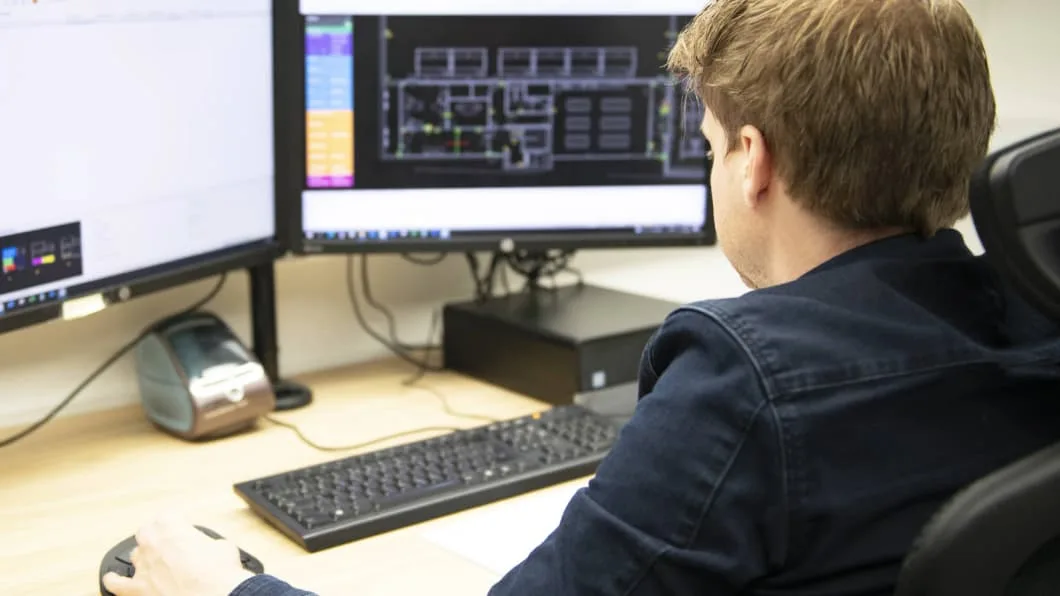 Person at a desk with dual monitors; the right screen displays a CAD floor plan. They are using an ergonomic mouse.