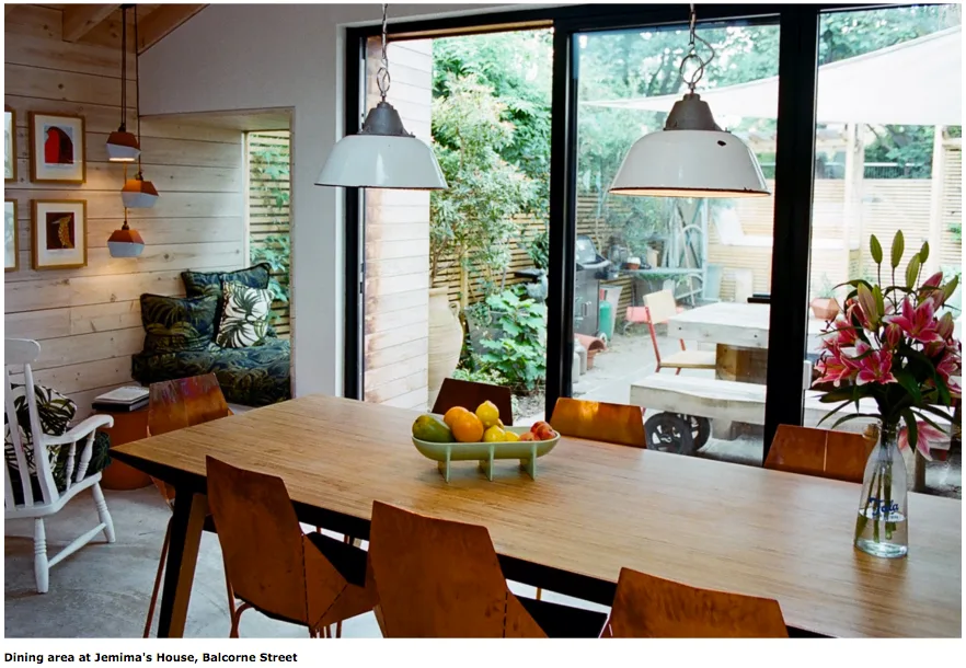 A wooden dining table is set with orange chairs and a fruit bowl, overlooking a garden patio.