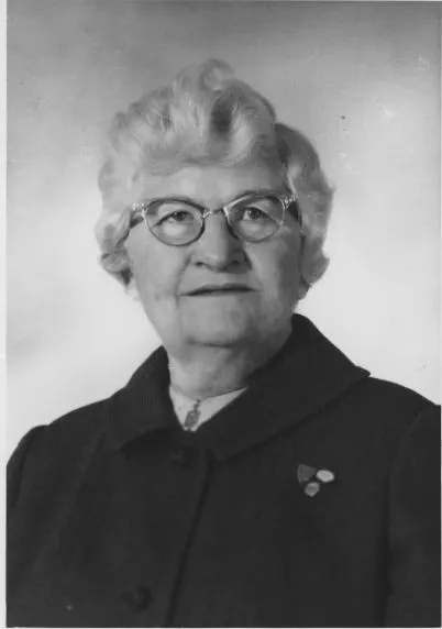 Portrait of a woman wearing glasses and a dark coat with a pin.