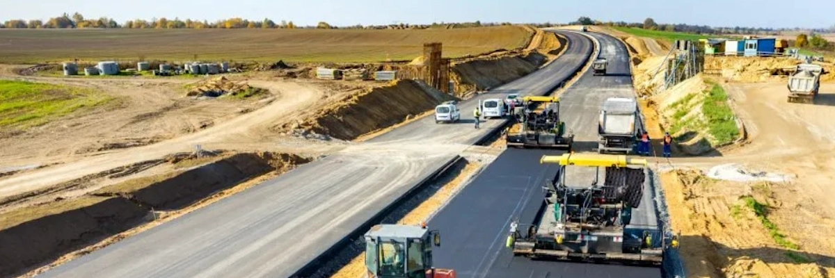 A construction site where a new road is being paved. Several construction vehicles, including asphalt pavers and rollers, are actively working on laying down the asphalt. Workers are visible near the vehicles.