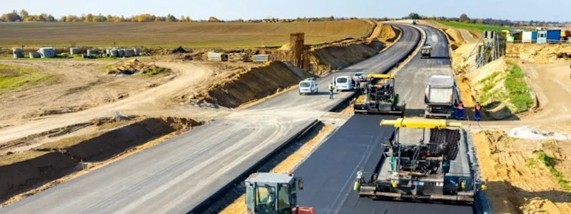 A construction site where a new road is being paved. Several construction vehicles, including asphalt pavers and rollers, are actively working on laying down the asphalt. Workers are visible near the vehicles.