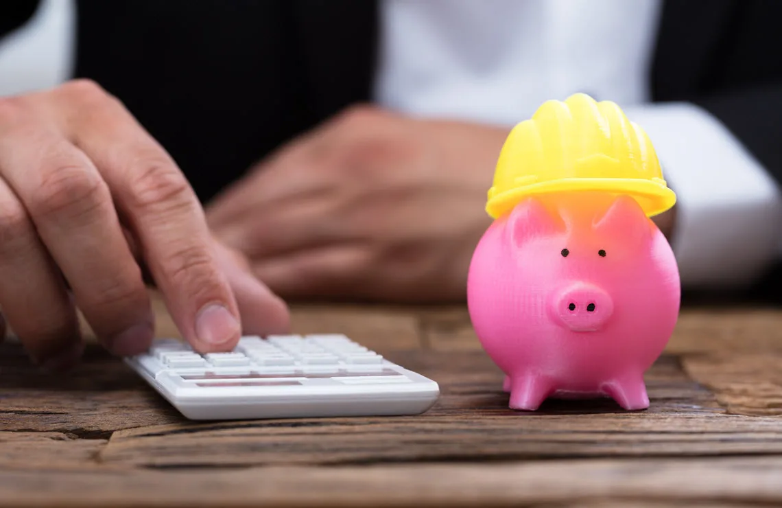 A person in a suit uses a calculator next to a pink piggy bank wearing a yellow hard hat.