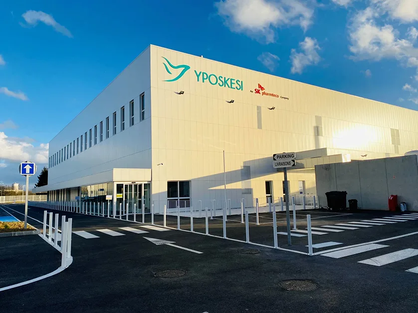 A modern, white industrial building with the YPOSKESI logo and SK pharmteco Company branding is shown.