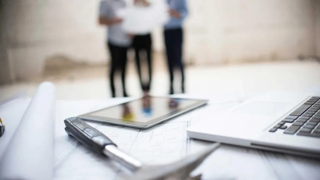 Close-up of a desk with a laptop, tablet and blueprints. A hammer lies on the plans with people discussing in the background.