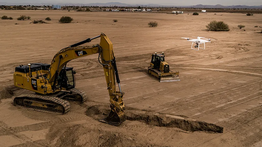 An excavator and bulldozer work on a construction site while a drone flies overhead.