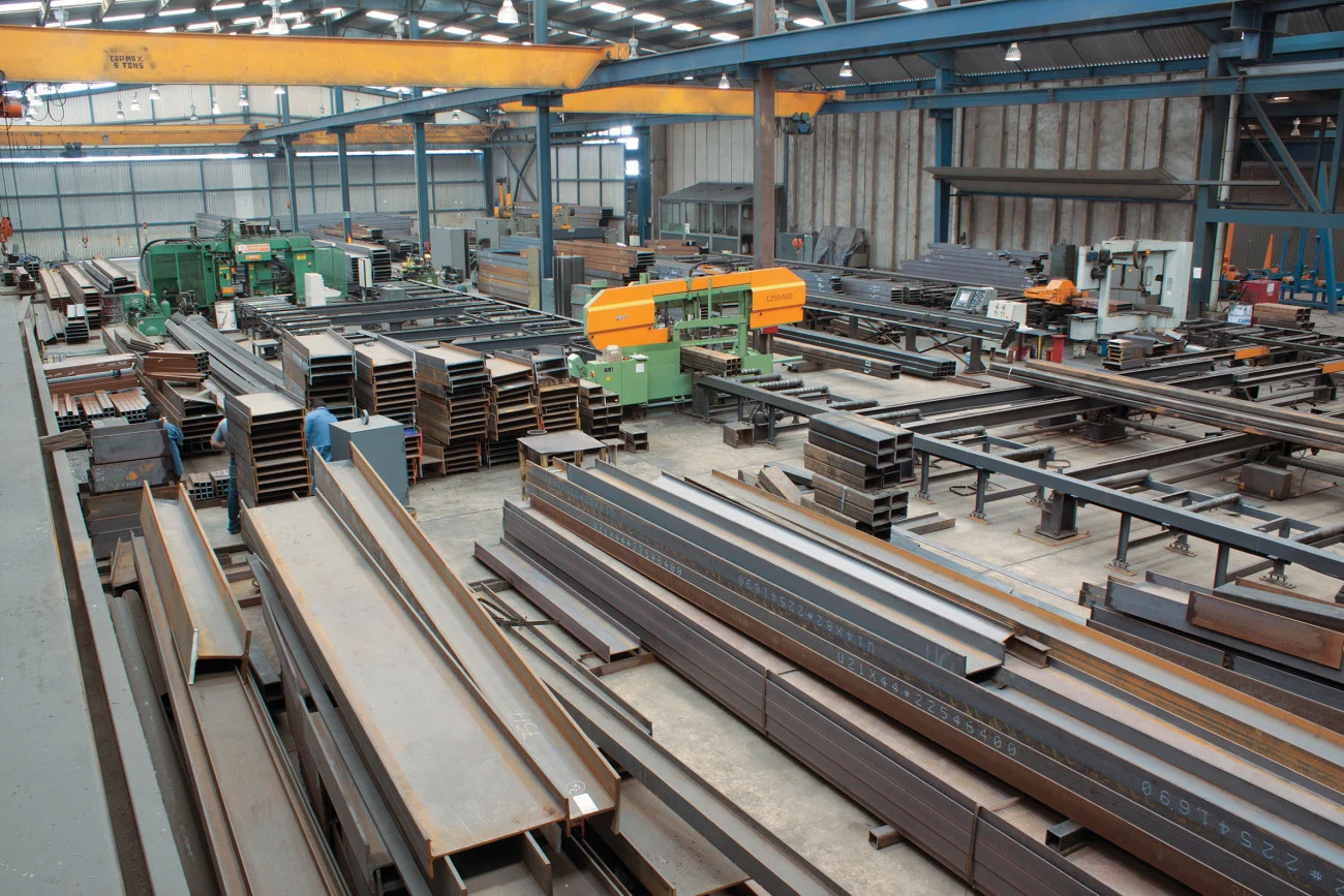 An overhead view of a metal fabrication workshop with stacks of steel beams and industrial machinery.