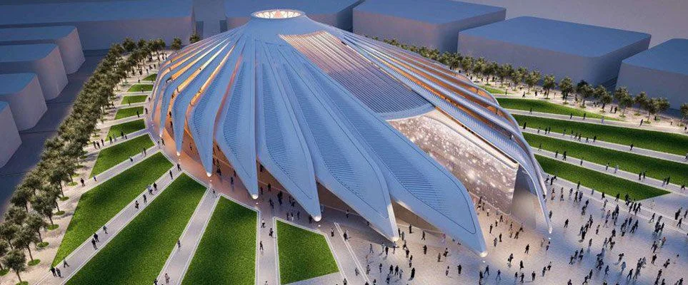An aerial view of a futuristic, petal-shaped building with large glass panels, surrounded by manicured lawns, walkways, and a crowd of people.