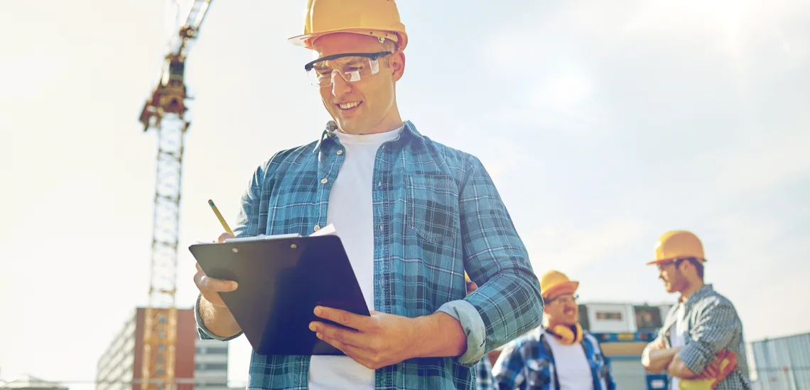 Construction worker in a hard hat holding a clipboard, with colleagues and a crane in the background on a sunny day