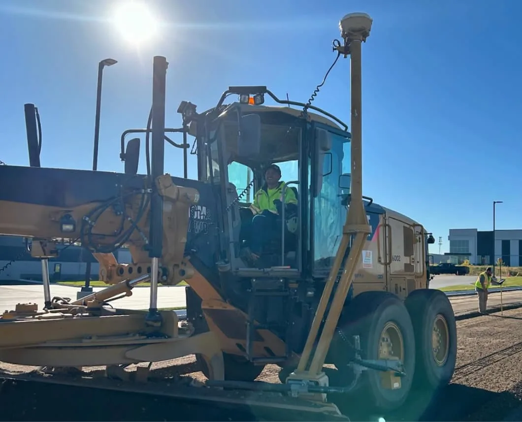 A construction worker in a yellow high-visibility vest operates a yellow grader with a GPS system on a construction site.