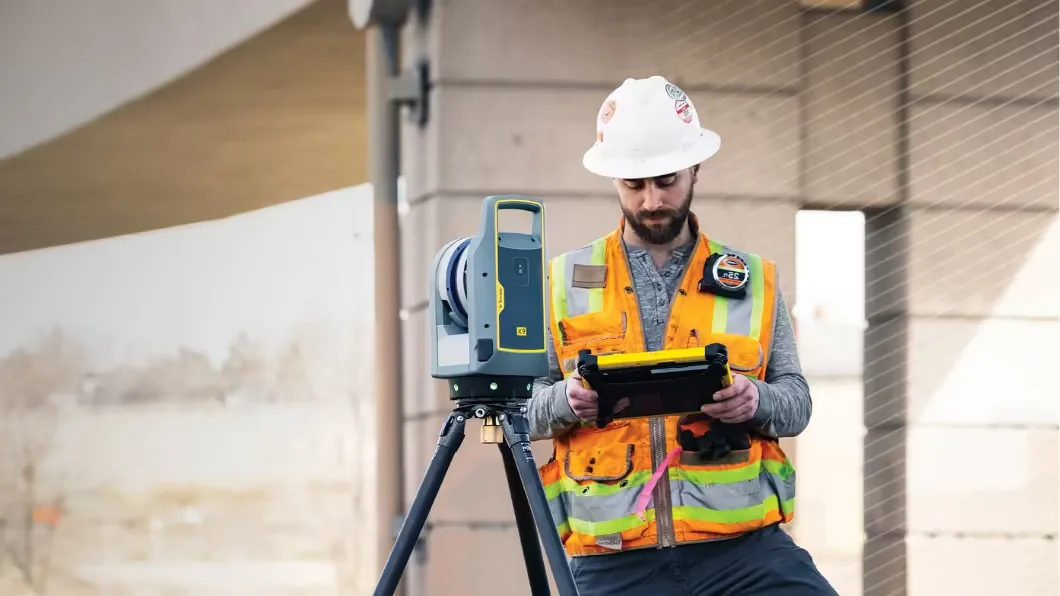 A construction worker in a hard hat and safety vest uses a tablet next to a Trimble X9 laser scanner.