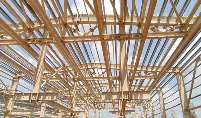 The steel framework of a large building under construction, showing multiple beams and supports against a cloudy sky.