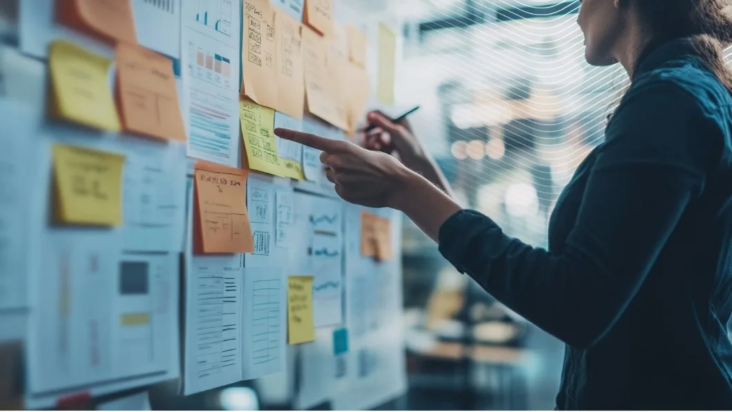A woman points at colorful sticky notes and documents on a planning board in an office setting.
