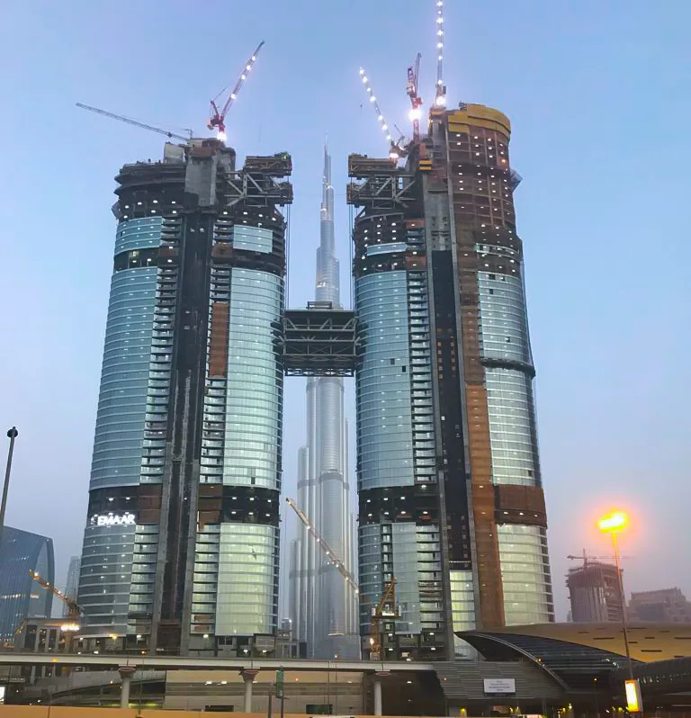 Two skyscrapers under construction are linked by a skybridge, with the Burj Khalifa visible in the background.