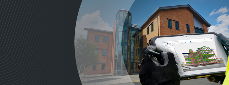 A person in a high-vis vest uses a tablet to view a 3D structural model in front of a modern brick building.