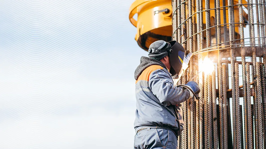 A construction worker in a welding mask assembles a large cylindrical rebar cage at an outdoor site with heavy machinery in the background.