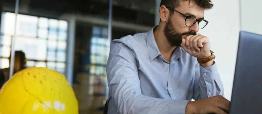A man with glasses and a beard looks intently at a laptop screen, with a yellow hard hat in the foreground.