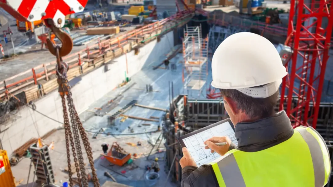 A person wearing a hardhat and safety vest stands above a large construction site and draws on a computer tablet with a stylus.